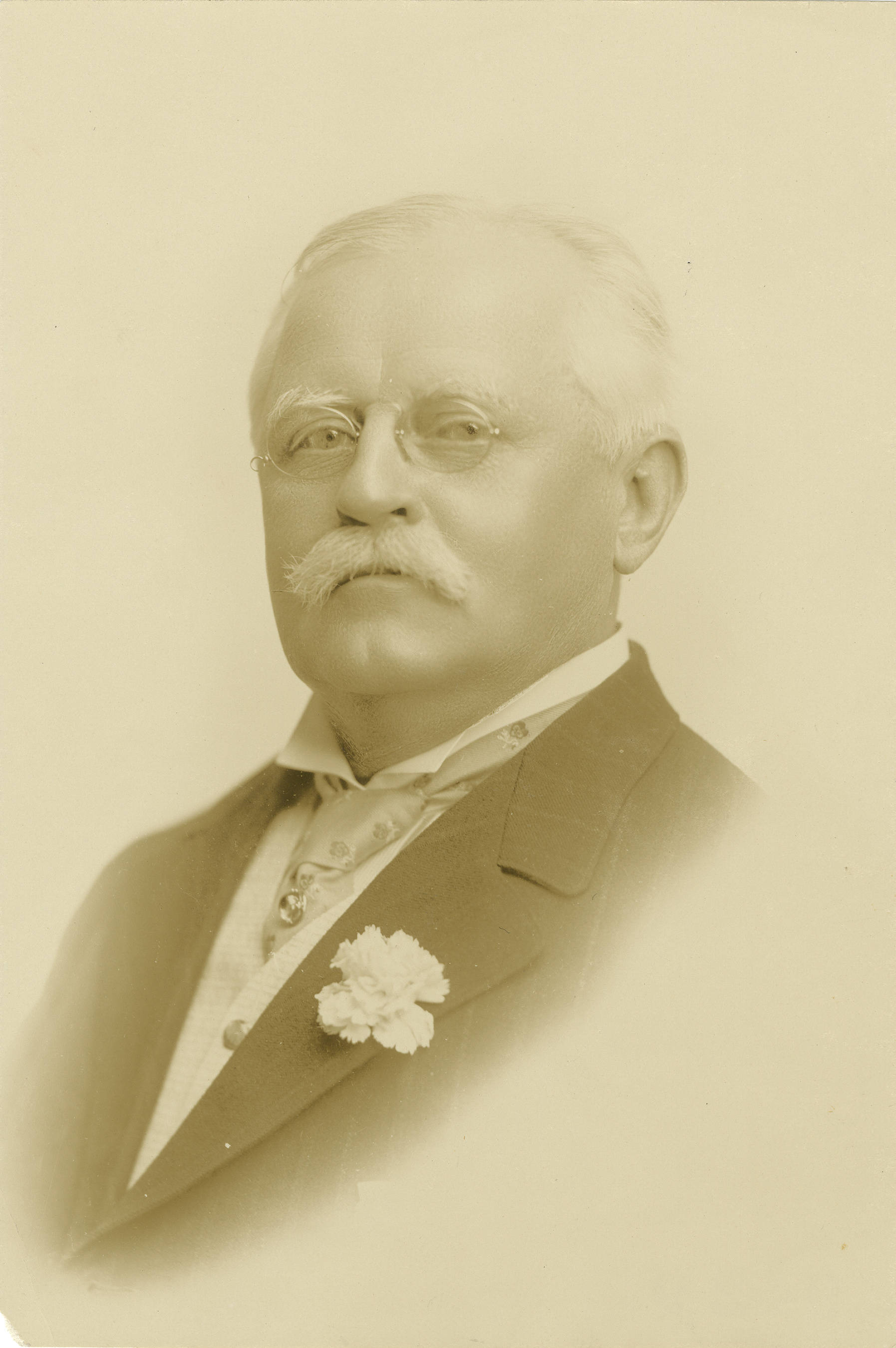 "A posed portrait of a man with short gray hair and mustache, wearing a jacket, necktie with pin, pince-nez eyeglasses, and a carnation on his jacket lapel, facing left and looking at the camera." Source: The Carolina Story.